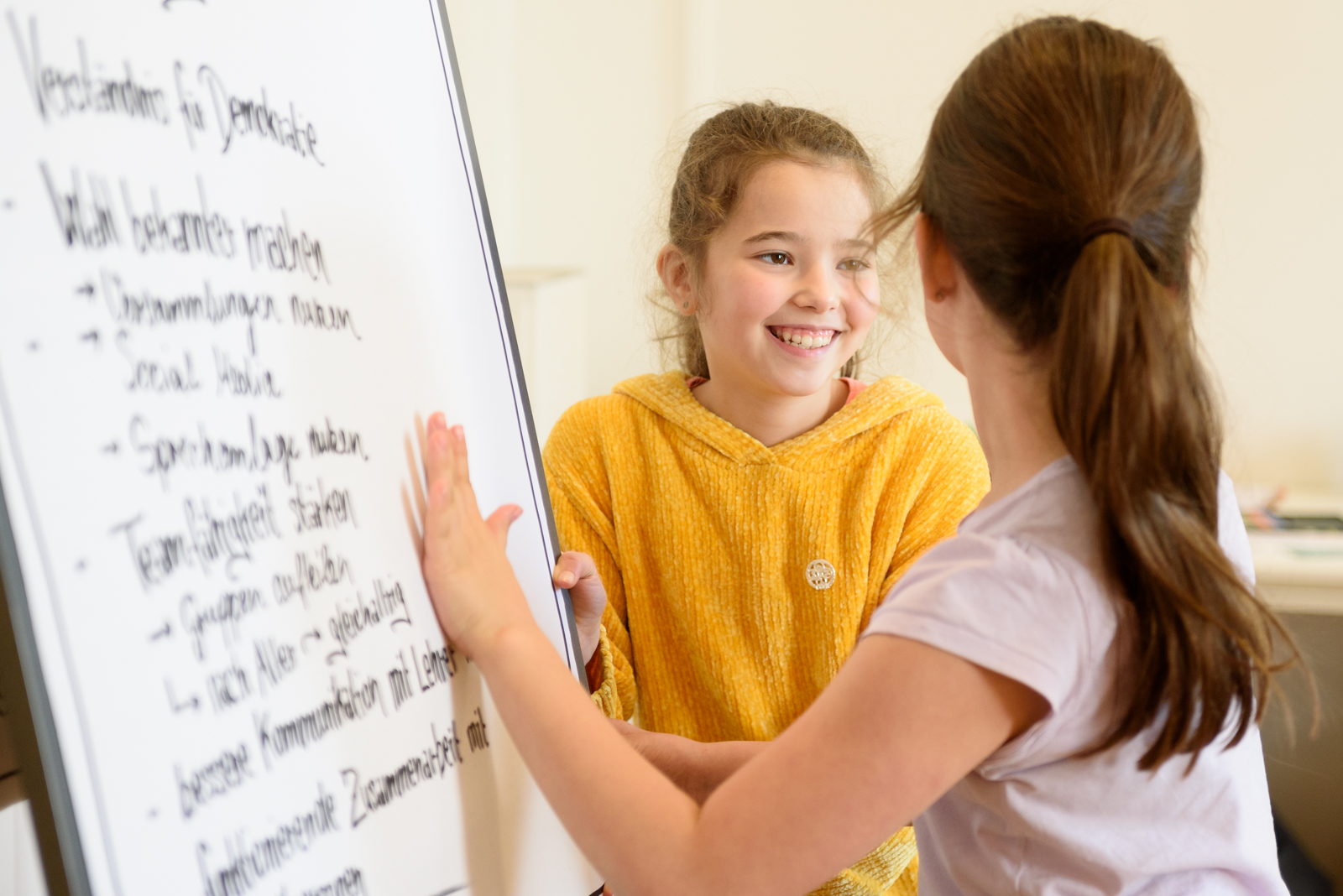 Zwei Mädchen stehen vor einem Flipchart und lächeln sich an.