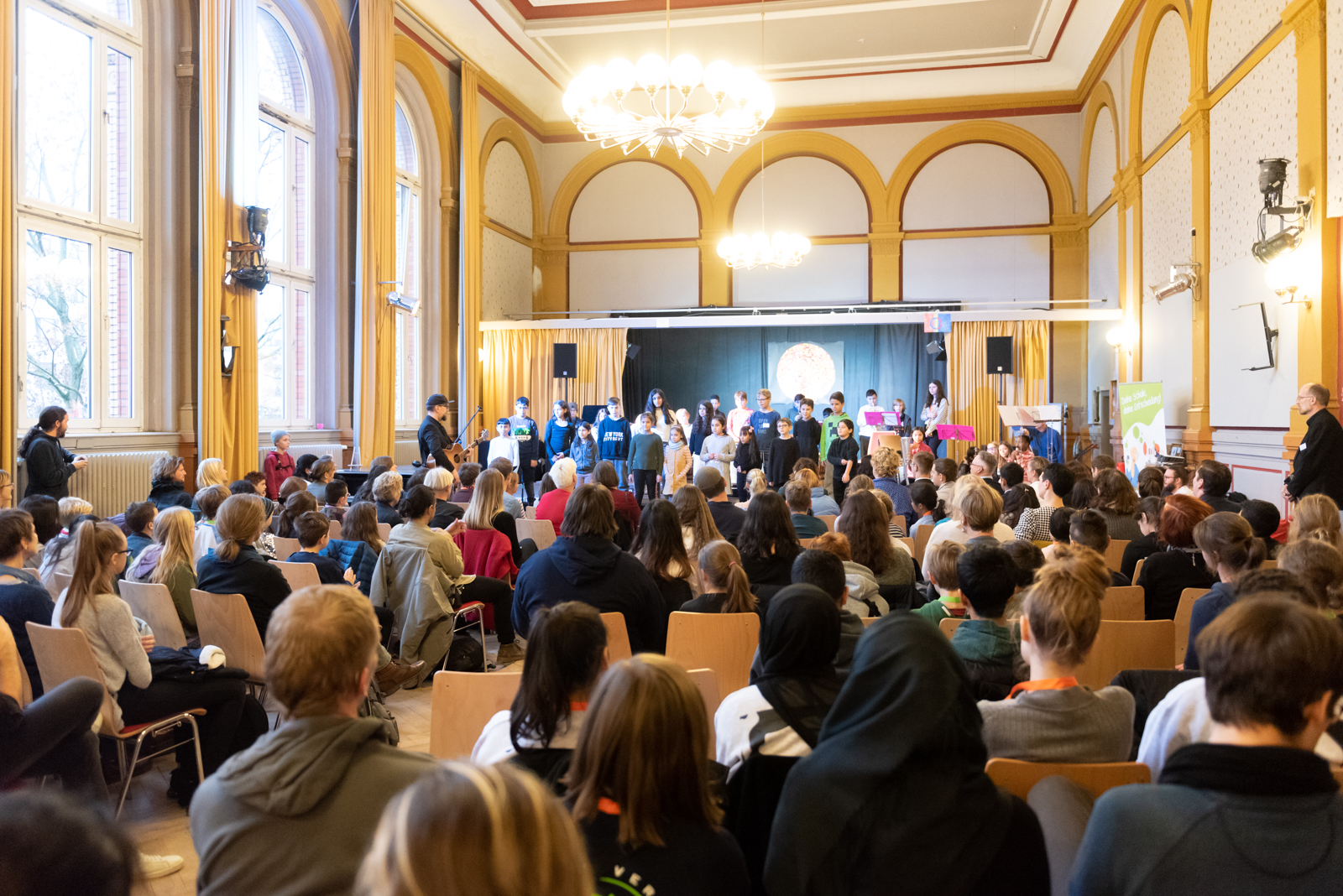 In einem Raum ist eine Bühne aufgebaut. Auf der Bühne steht eine Gruppe von Schüler*innen. Vor der Bühne sitzen viele Zuschauer im Plenum.