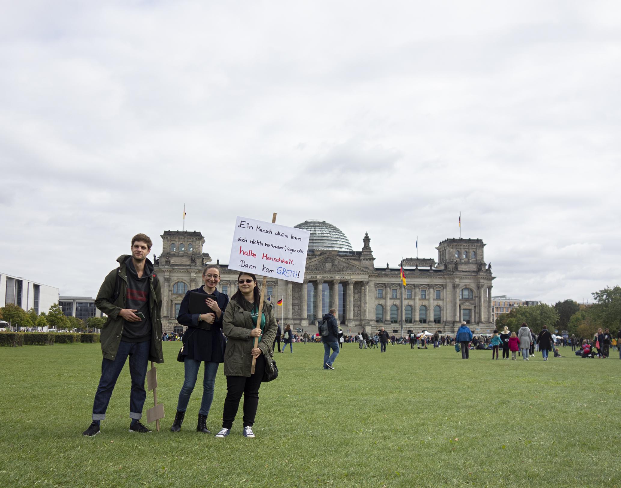 Drei Personen stehen auf der Wiese vor dem Reichstagsgebäude. Eine der Personen hat ein Schild in der Hand. Auf dem Schild steht: "Ein Mensch alleine kann doch nichts verändern. sagte die halbe Menschheit. Dann kam Greta."