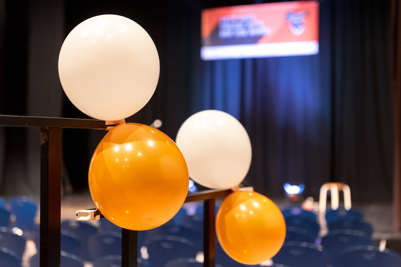 In einer großen Aula ist vorne eine Bühne und davor wurden Stühle aufgebaut. Im Fokus sind vier Luftballons zu sehen. Jeweils zwei der Ballons, ein weißer und ein orangener hängen zusammen an einem Geländer.