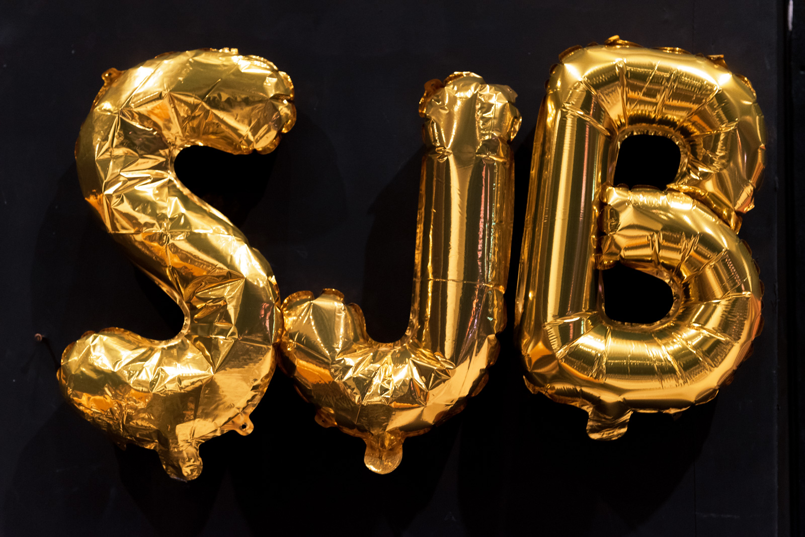 Drei goldene Folienballons sind vor einem schwarzen Hintergrund. Die Folienballons sind Buchstaben und stellen das Wort "SJB" da.