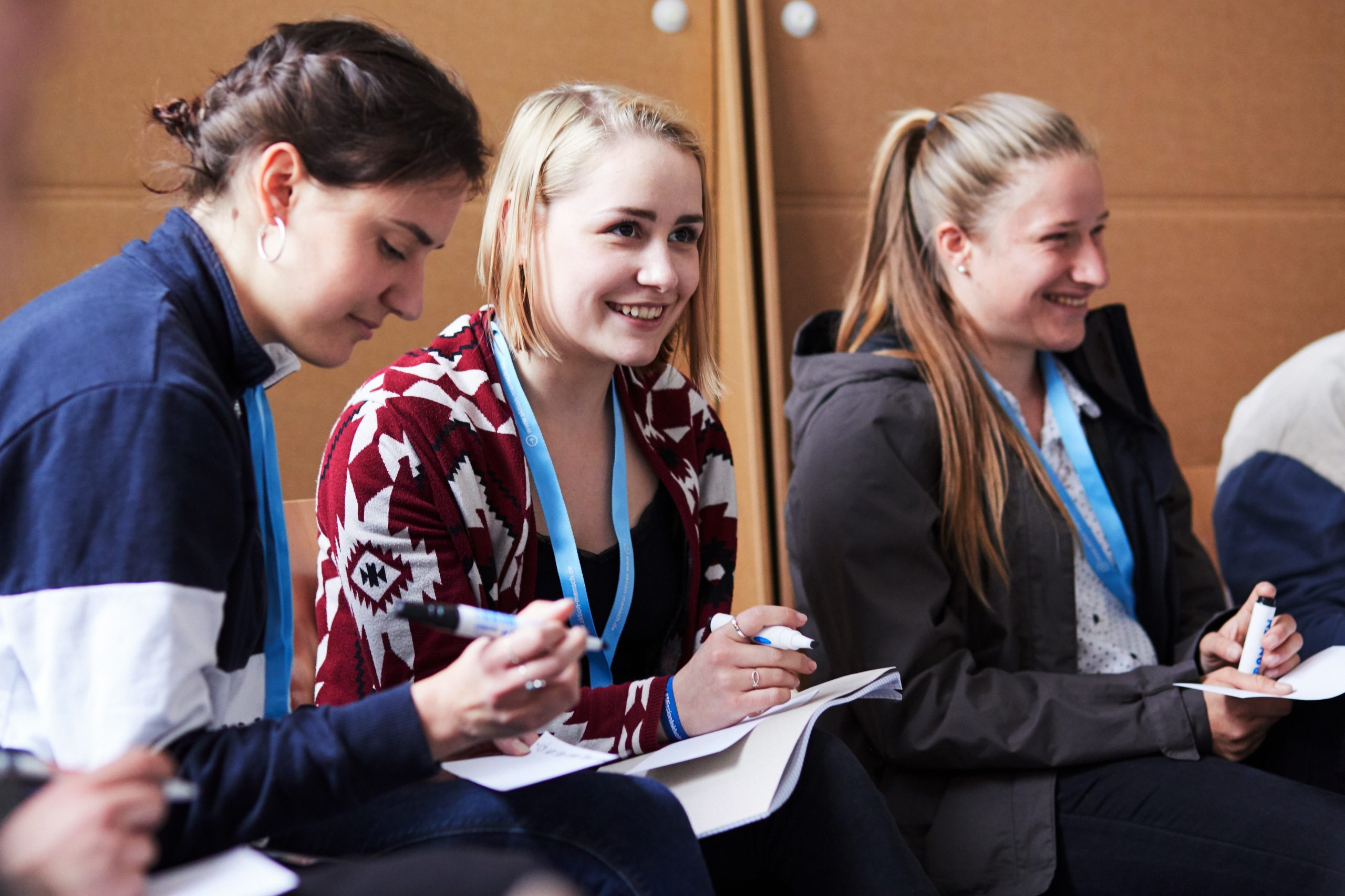 Mission Inklusion — Jugendaktionscamp 2019 Drei junge Frauen sitzen in einer Reihe und lächeln. Sie haben einen Block auf dem Schoß und einen Stift in der Hand.