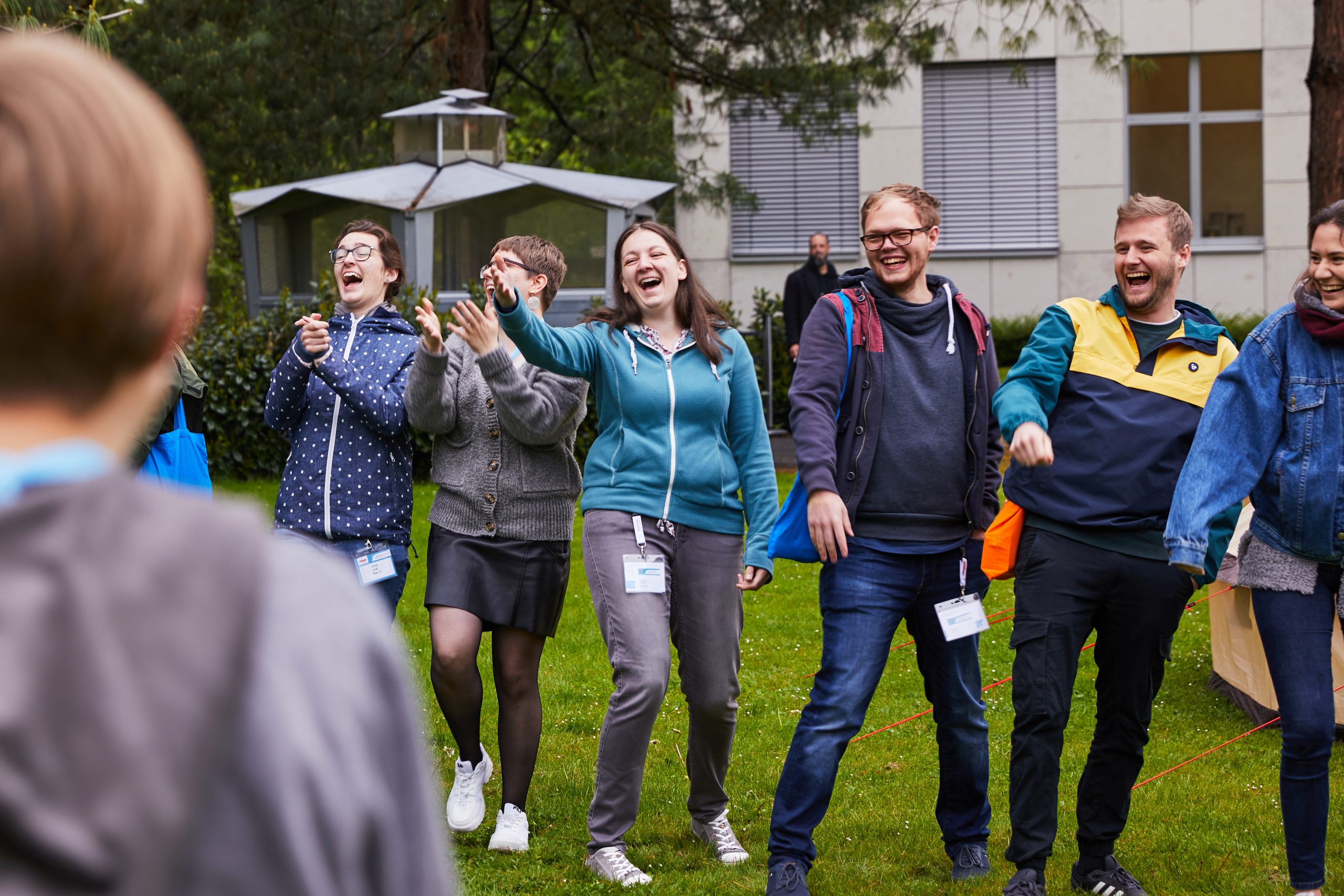 Eine Gruppe junger Menschen steht in einer Reihe auf einem Rasen. Ihnen gegenüber stehen weitere Personen. Die Gruppe lacht und applaudiert.