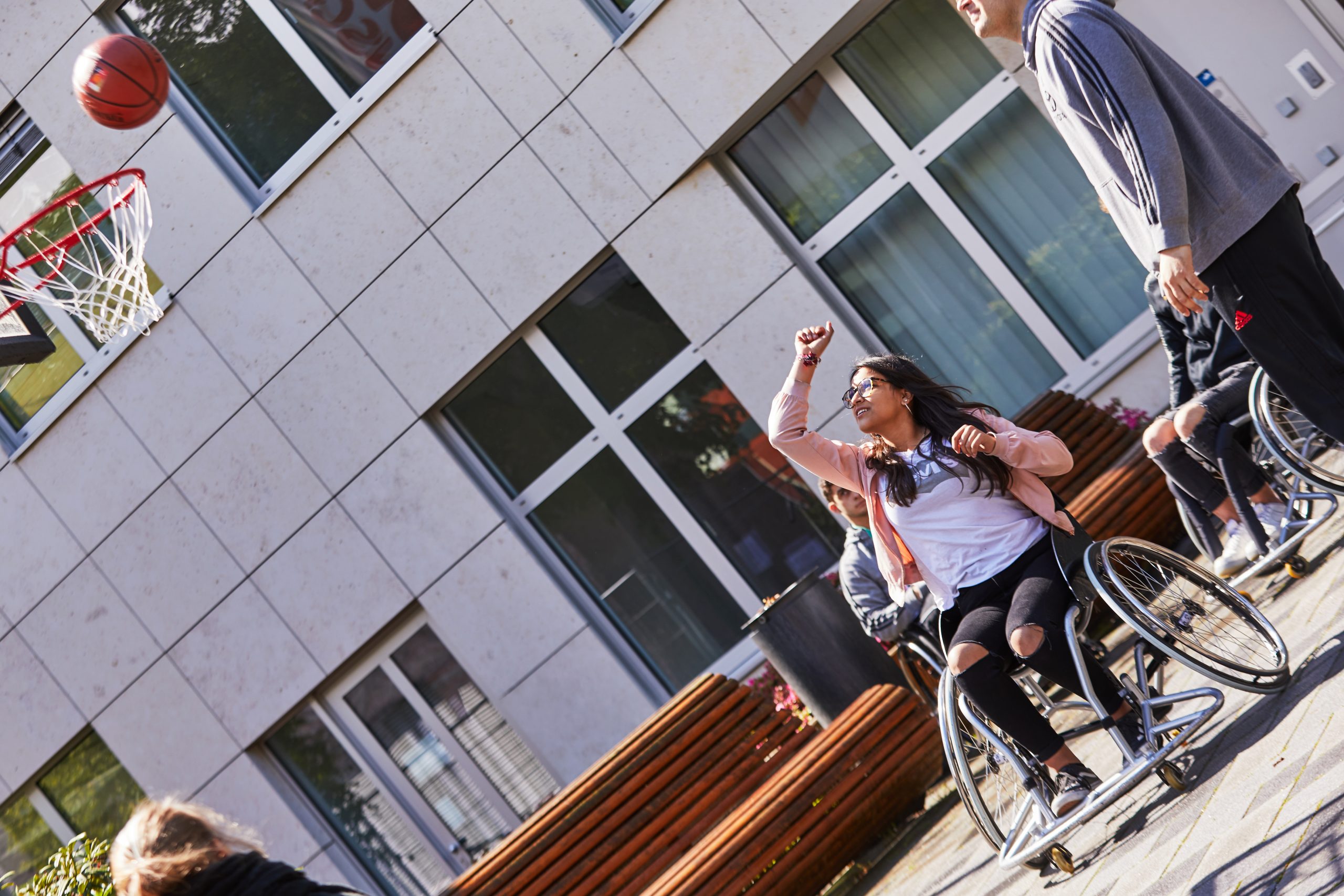 Eine junge Frau sitzt in einem Rollstuhl und wirft mit einem Basketball auf einen Basketballkorb. Neben ihr steht ein junger Mann.