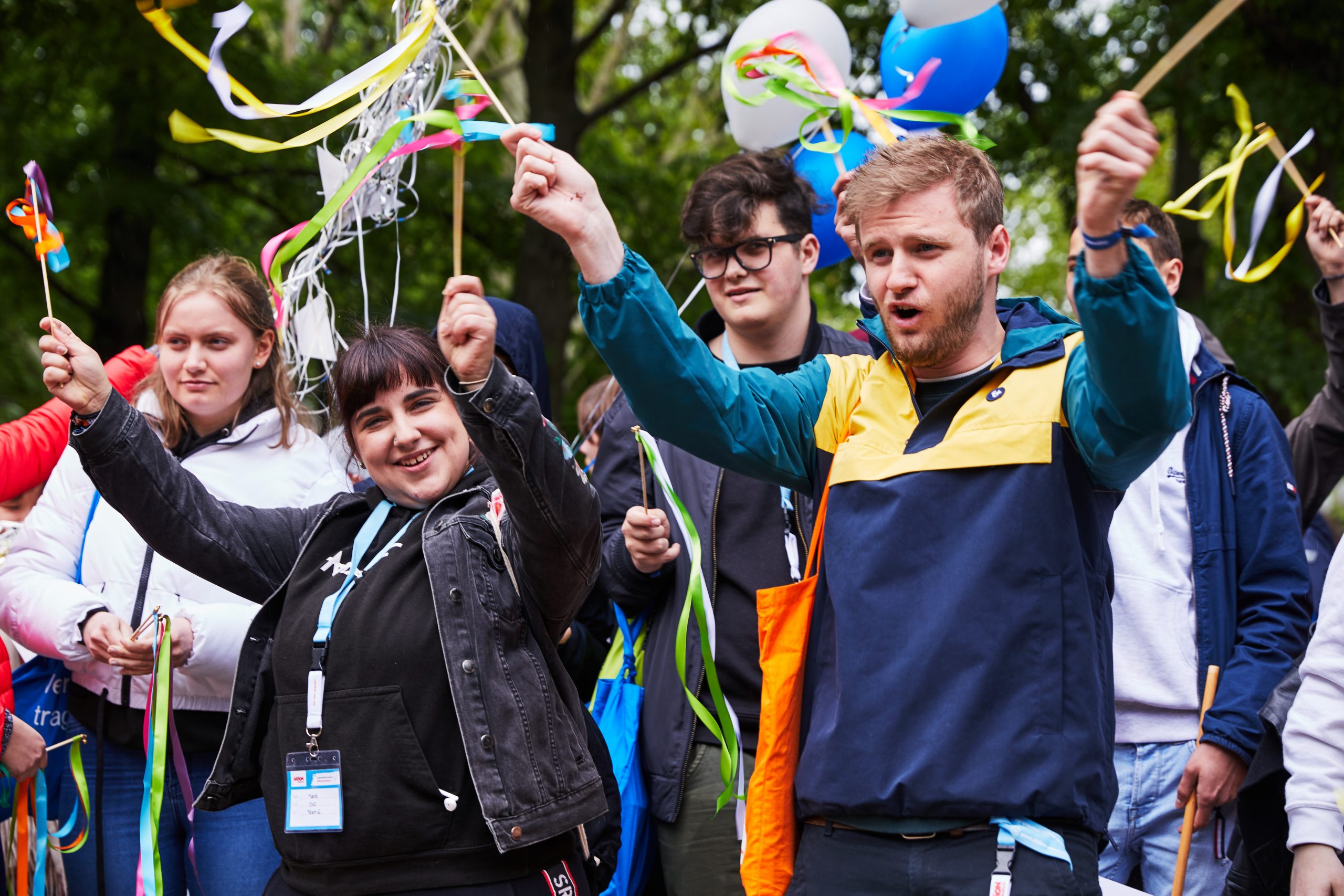 Eine Gruppe von jungen Menschen hält Stäbe mit bunten Bändern und Luftballons in der Hand. Die Gruppe läuft draußen und sieht glücklich aus.