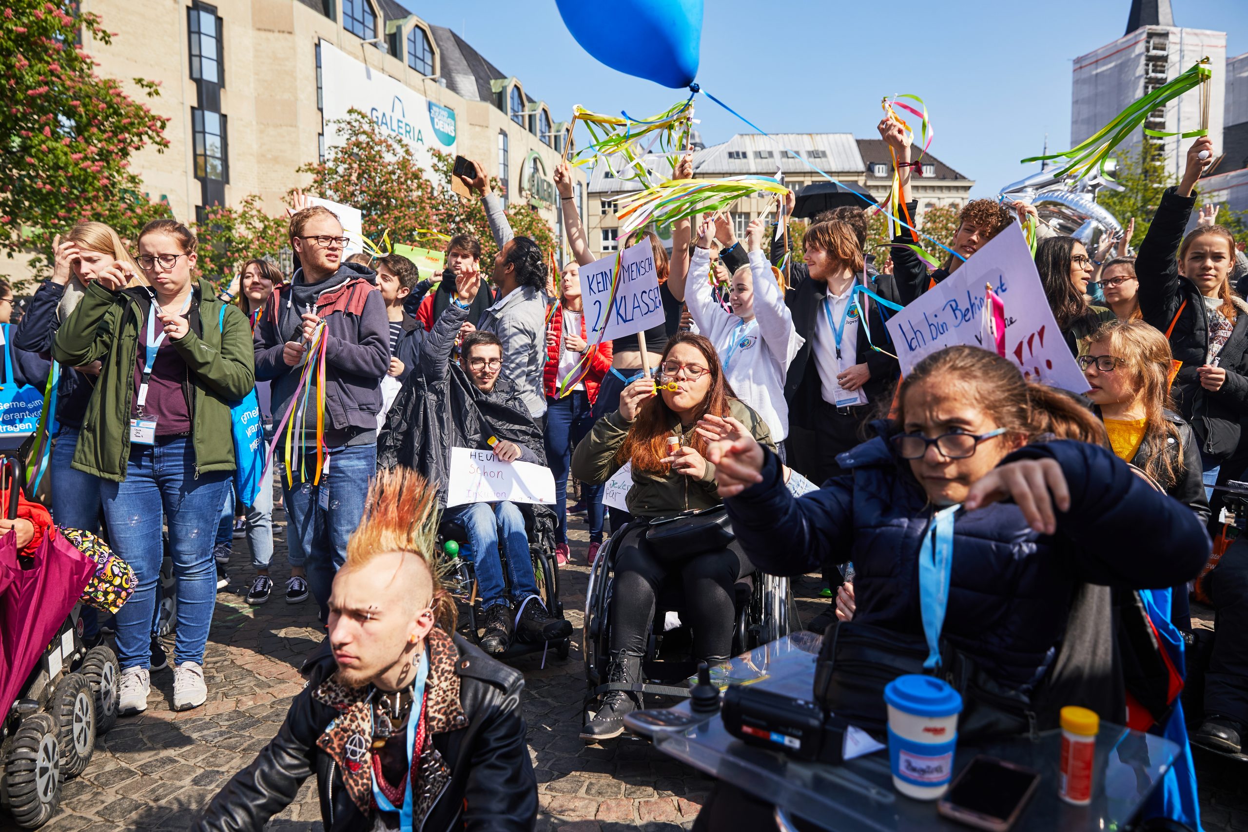 Viele unterschiedlich junge Menschen mit und ohne sichtbare Behinderung stehen in einer Gruppe. Einige Personen halten Plakate in die Luft. Andere haben Luftballons oder bunte Bänder in der Hand.