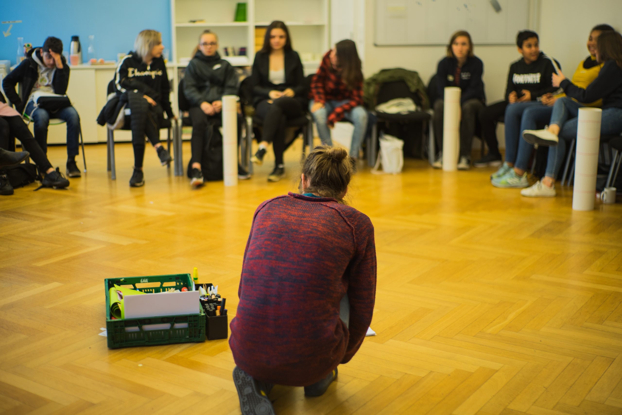 Auf dem Bild sieht man verschiedene Menschen, die in einem Stuhklreis sitzen. In der Mitte kniet sich eine Person, um Moderationsmaterial für den nächsten inhaltlichen Block aufzuheben. © Servicestelle Jugendbeteiligung e.V., 2019