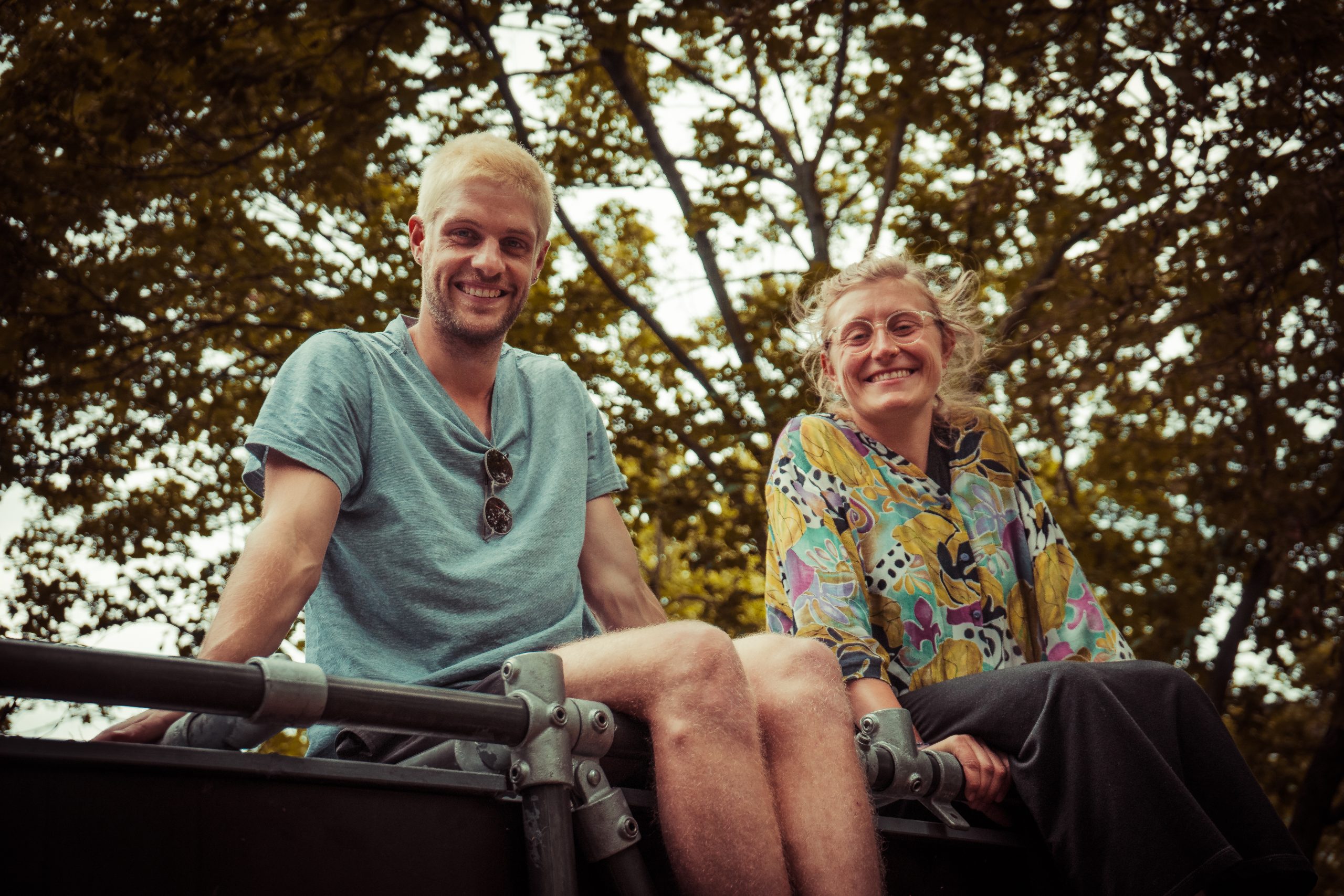 Ein junger Mann und eine junge Frau sitzen lachend auf einem Gerüst. Im Hintergrund sind Bäume zu sehen.