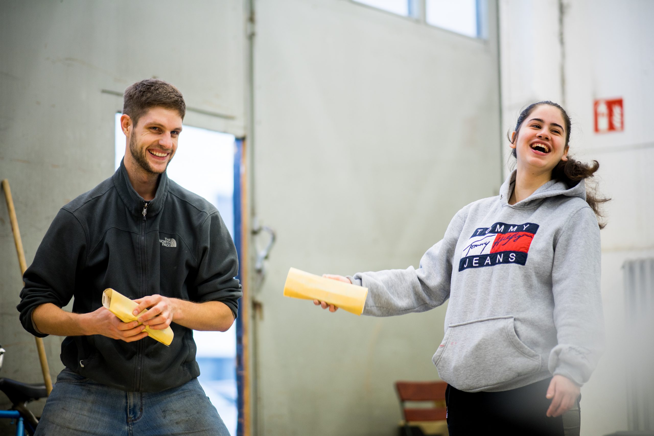 Ein junger Mann und eine junge Frau stehen lachend in einer Halle. Die beiden halten Schleifpapier in der Hand.