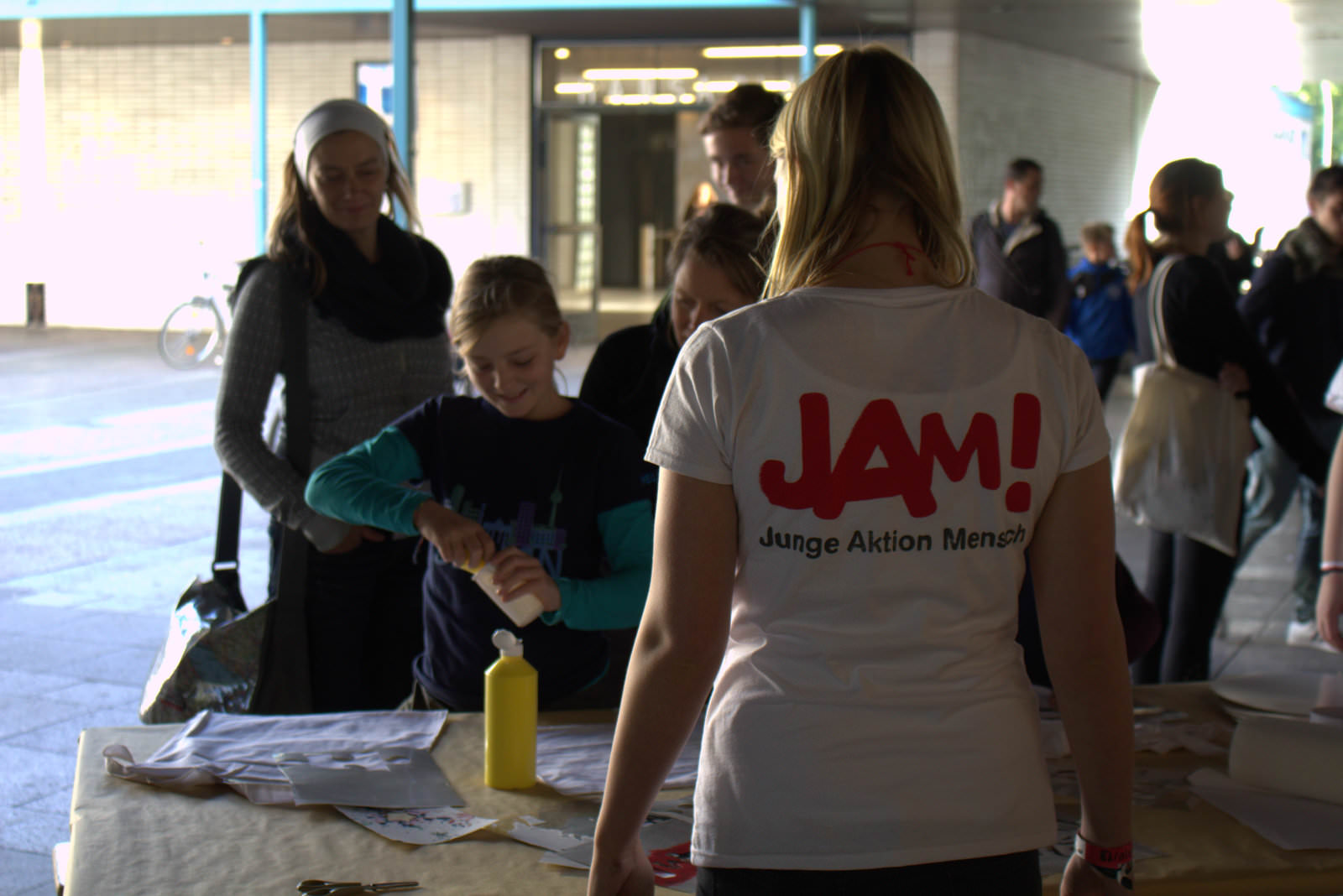 Eine Person steht mit dem Rücken zur Kamera. Sie trägt ein weißes Tshirt mit der Aufschrift JAM. Vor ihr steht ein Tisch und mehrere Menschen, die Taschen bemalen.