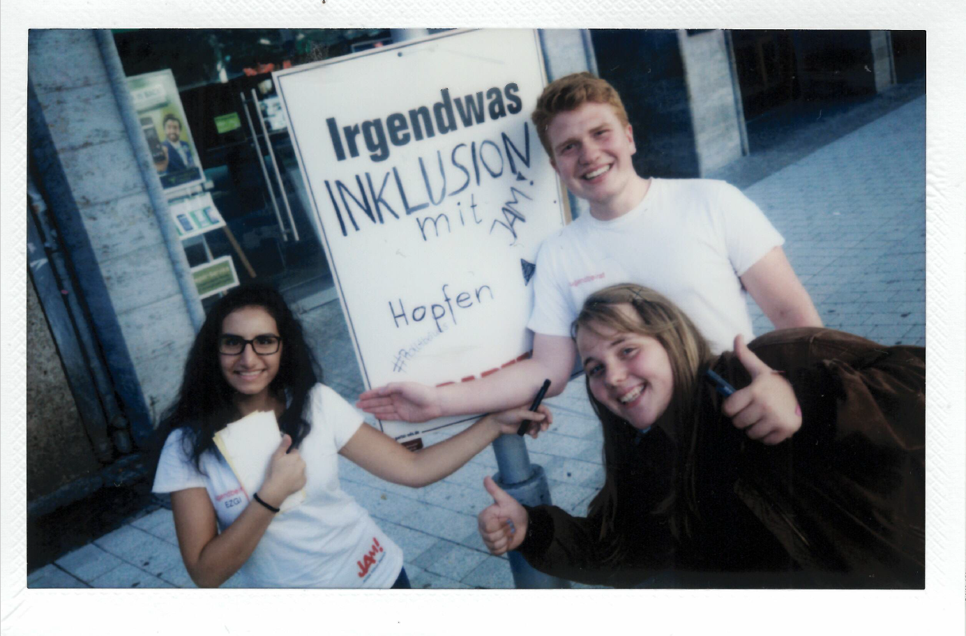 Zwei junge Frauen und ein junger Mann stehen vor einem Schild, lachen und halten die Daumen nach oben. Auf dem Schild steht: Irgendwas mit Inklusion.