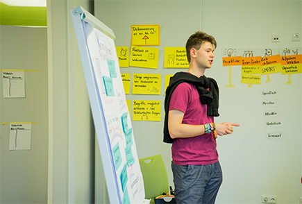 Das Bild zeigt einen jungen Mann, der vor einem Flipchart steht und etwas erklärt. Im Hintergrund sind viele Moderationskarten an die Wand gepinnt. © Servicestelle Jugendbeteiligung e.V., 2019