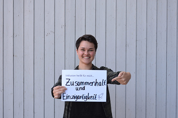 Das Bild zeigt einen jungen Menschen, der vor einer weißen Wand steht. Sie hält ein Schild in der Hand, auf dem steht "Inklusion heißt für mich, Zusammenhalt und Einzigartigkeit" © Servicestelle Jugendbeteiligung e.V., 2018