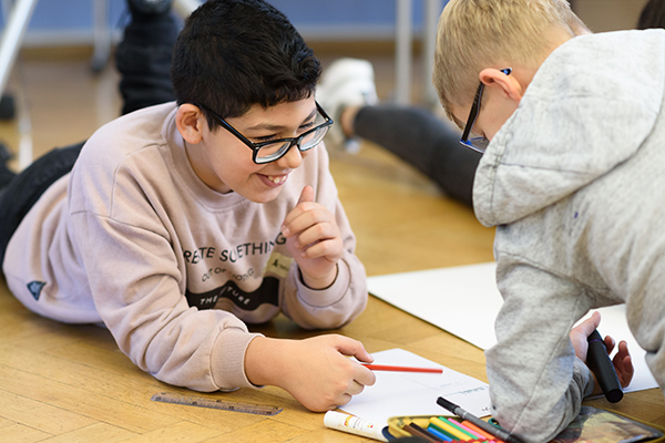 Auf dem Bild sieht man zwei Personen auf dem Boden liegen und mit Buntstiften auf einem Blatt malen. Die eine Person lächelt. © Servicestelle Jugendbeteiligung e. V. 2019