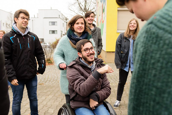 Im Fokus ist ein junger Mann im Rollstuhl zu sehen. Er wird von einer jungen Frau geschoben. Im Hintergrund sind weitere junge Menschen zu erkennen. © Servicestelle Jugendbeteiligung e.V., 2016