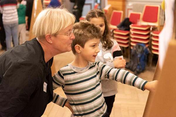 Zwei Kinder stehen mit ihrer Lehrerin vor einer Pinnwand und diskutieren über den Inhalt auf der Pinnwand. Ein Kind zeigt auf etwas. © Servicestelle Jugendbeteiligung e.V., 2020