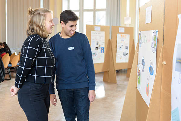 Auf dem Bild sieht man zwei Personen, die sich Plakate anschauen, die auf Pinnwänden aus Pappe angebracht sind. Im Hintergrund sieht man mehr Pinnwände. © Servicestelle Jugendbeteiligung e.V., 2020