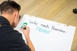 Das Bild zeigt eine Person, die am Boden liegt und ein Flipchart mit der Überschrift "Auf der Suche nach Barrierefreiheit" gestaltet.© Servicestelle Jugendbeteiligung e. V., 2020