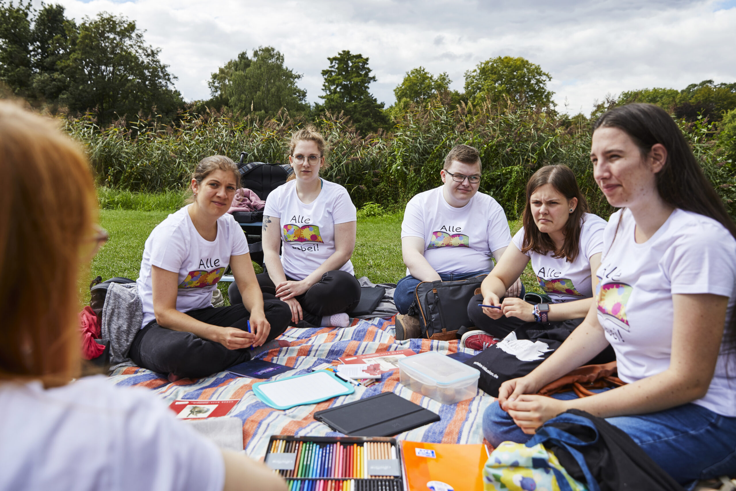 Das Bild zeigt fünf junge Menschen mit und ohne Behinderung, die auf einer Decke auf einer Wiese sitzen. Alle tragen T-Shirts mit der Aufschrift "Alle dabei" © Servicestelle Jugendbeteiligung / Aktion Mensch I Anna Spindelndreier