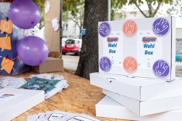 Das Bild zeigt einen Teil des U18-Wahlstandes von JMP. Auf dem Holztisch sind, neben Luftballons und Broschüren, bunte Boxen mit der Aufschrift "SuperWahlBox" aufgetürmt.© Rainer Kurzeder, 2021