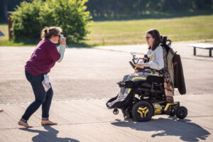 B: Eine Teilnehmende im Rollstuhl wird fotografiert. Die Person die das Foto macht und die Person die fotografiert wird scheinen Spaß dabei zu haben und lachen beide.