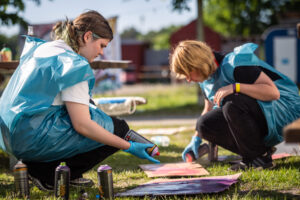 B: Zwei Teilnehmende knien über Leinwände und besprühen diese mit Farbe. beide tragen blaue Müllsäcke, als Schutz für die Kleidung.