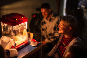 B: Eine beleuchtete Popcornmaschine, im dunklen Raum: Zwei Menschen knien davor und strahlen, breit lächelnd die Popcornmaschine an.