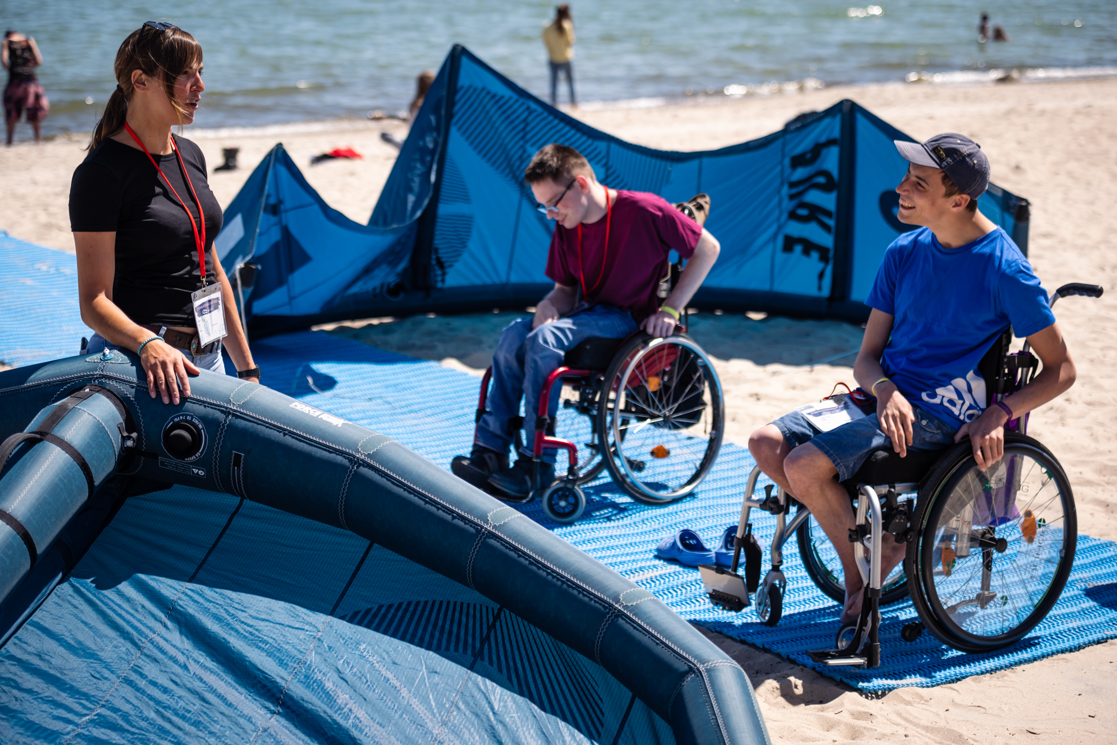 Das Bild zeigt zwei junge Menschen, die einen Rollstuhl nutzen beim Aufbau eines Kit-Drachens am Strand.