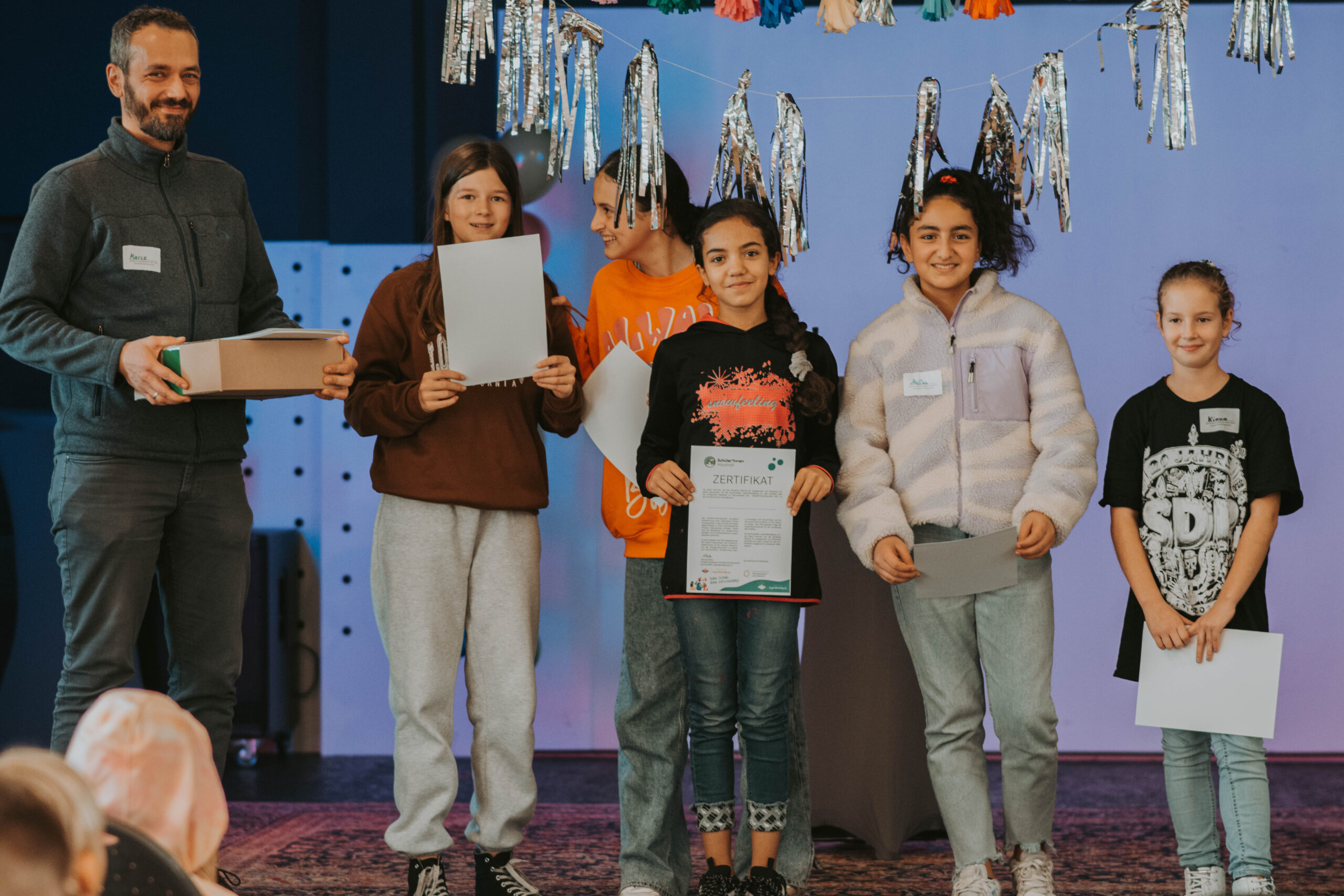 Das Bild zeigt Kinder und eine erwachsene Begleitperson, die auf einer Bühne Zertifikate überreicht bekommen ©Servicestelle Jugendbeteiligung 2022