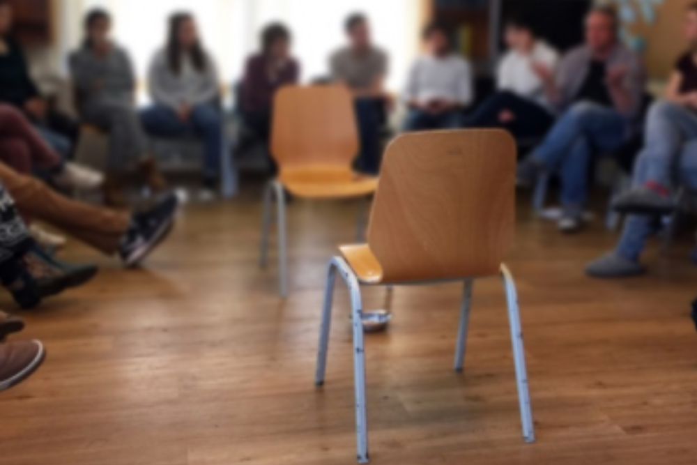 Das Bild zeigt einen Stuhl. Außerdem sitzen Jugendliche in einem Stuhlkreis um den Stuhl herum. Servicestelle Jugendbeteiligung 2023 ©
