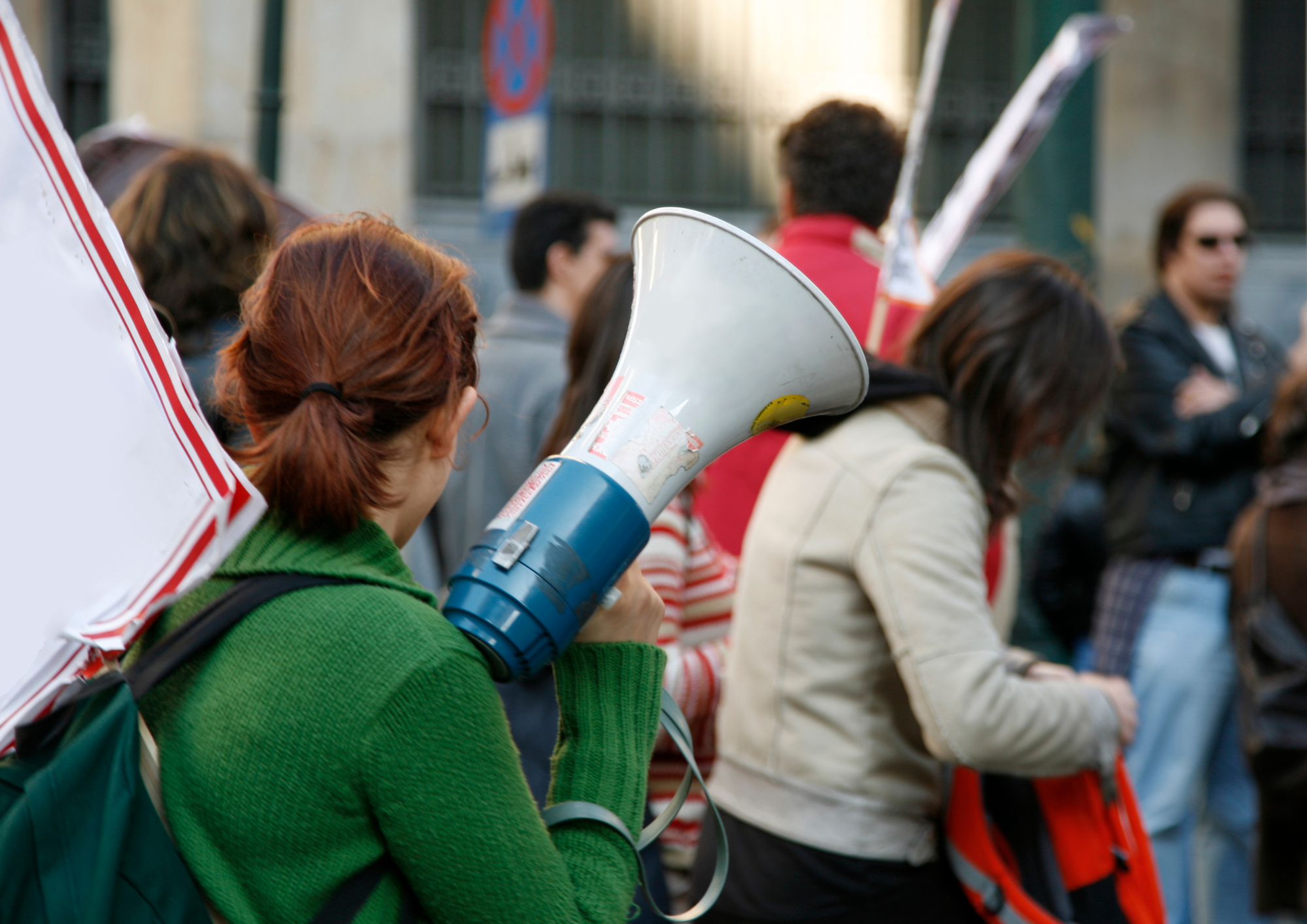 Person im Vordergrund mit einem weißen Schild auf der Schulter und einem Megafon in der Hand. Im Hintergrund sieht man weitere Personen