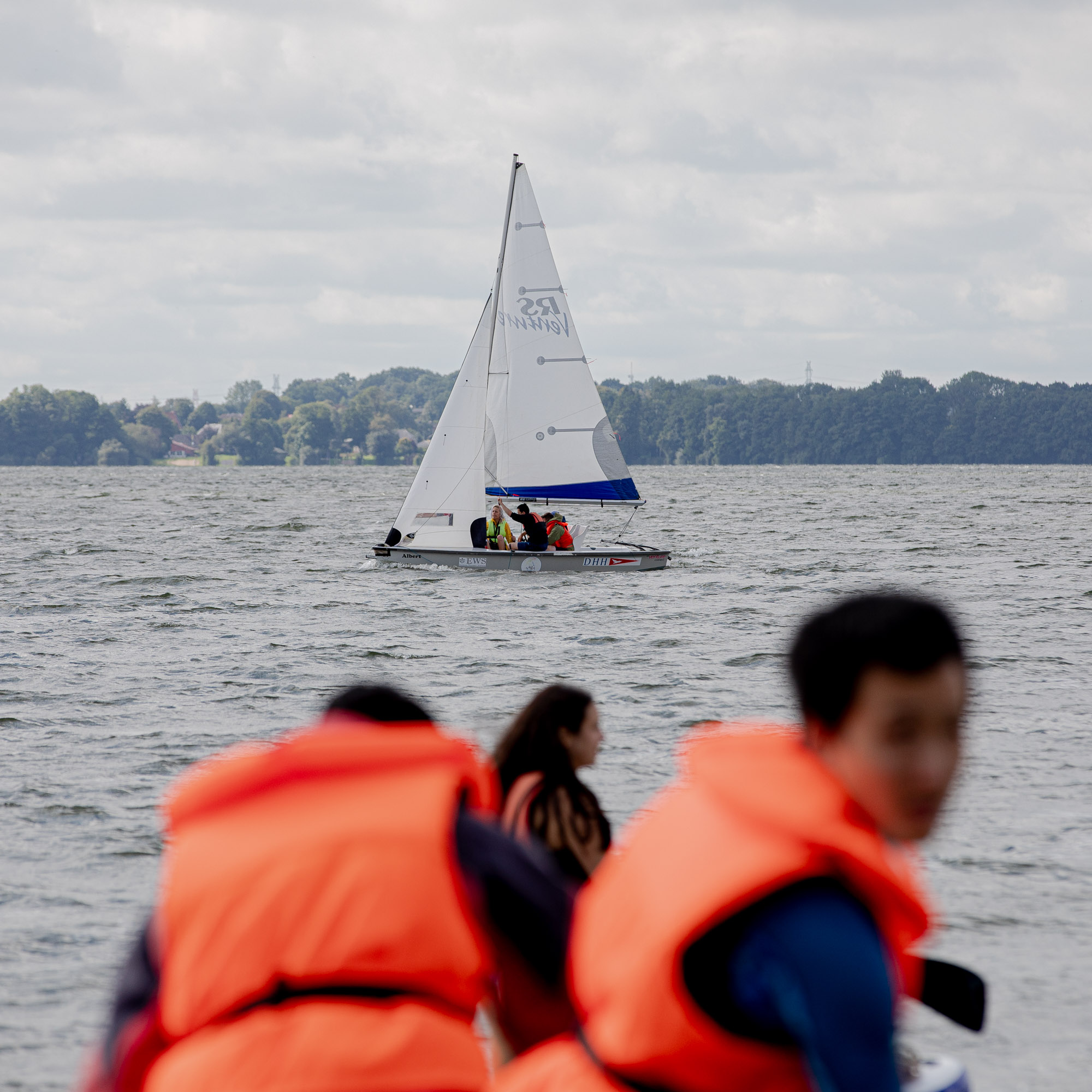 Im Vordergrund sieht man unscharf zwei Personen mit orangen Rettungswesten. Eine Person blick Richtung Kamera. Im Hintergrund sieht man ein Segelboot mit einem weißen Segel auf einem See. Auf dem Segelboot sind Menschen zu sehen.