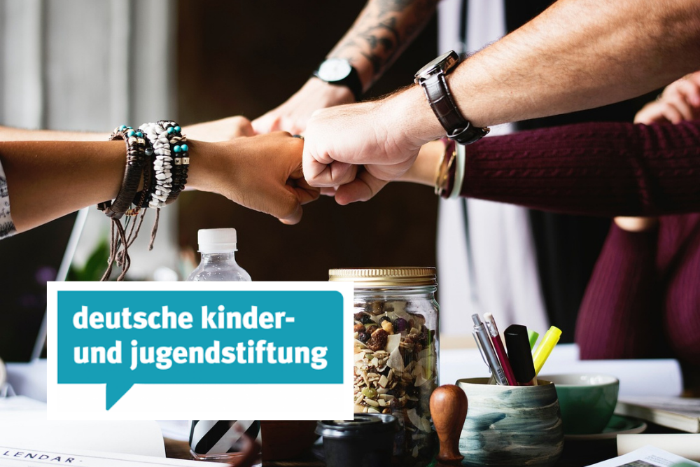 Im Hintergrund treffen sich mehrere Fäuste, über einem Schreibtisch als Arbeitsfläche, in der Mitte als Zeichen der Zusammenarbeit. Im Vordergrund ist das Logo der deutschen kinder- und jugendstiftung abgebildet.