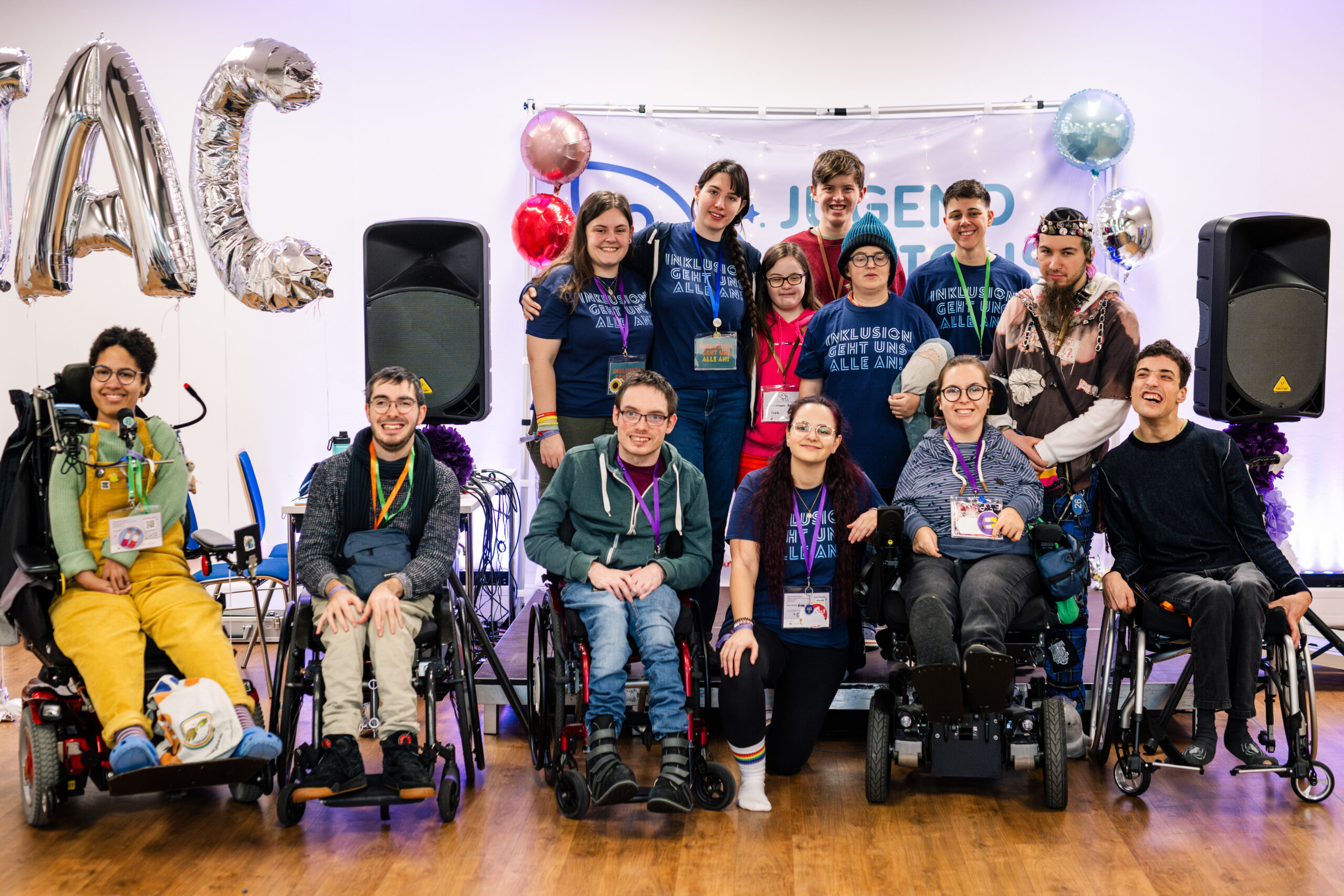 Das Bild zeigt eine Gruppe von Menschen auf und vor einer Bühne. Es sind Menschen im Rollstuhl zu sehen, Menschen die knien und Menschen die stehen. Im Hintergrund ist ein Banner zu sehen mit der Aufschrift Jugendaktionscamp 2023.