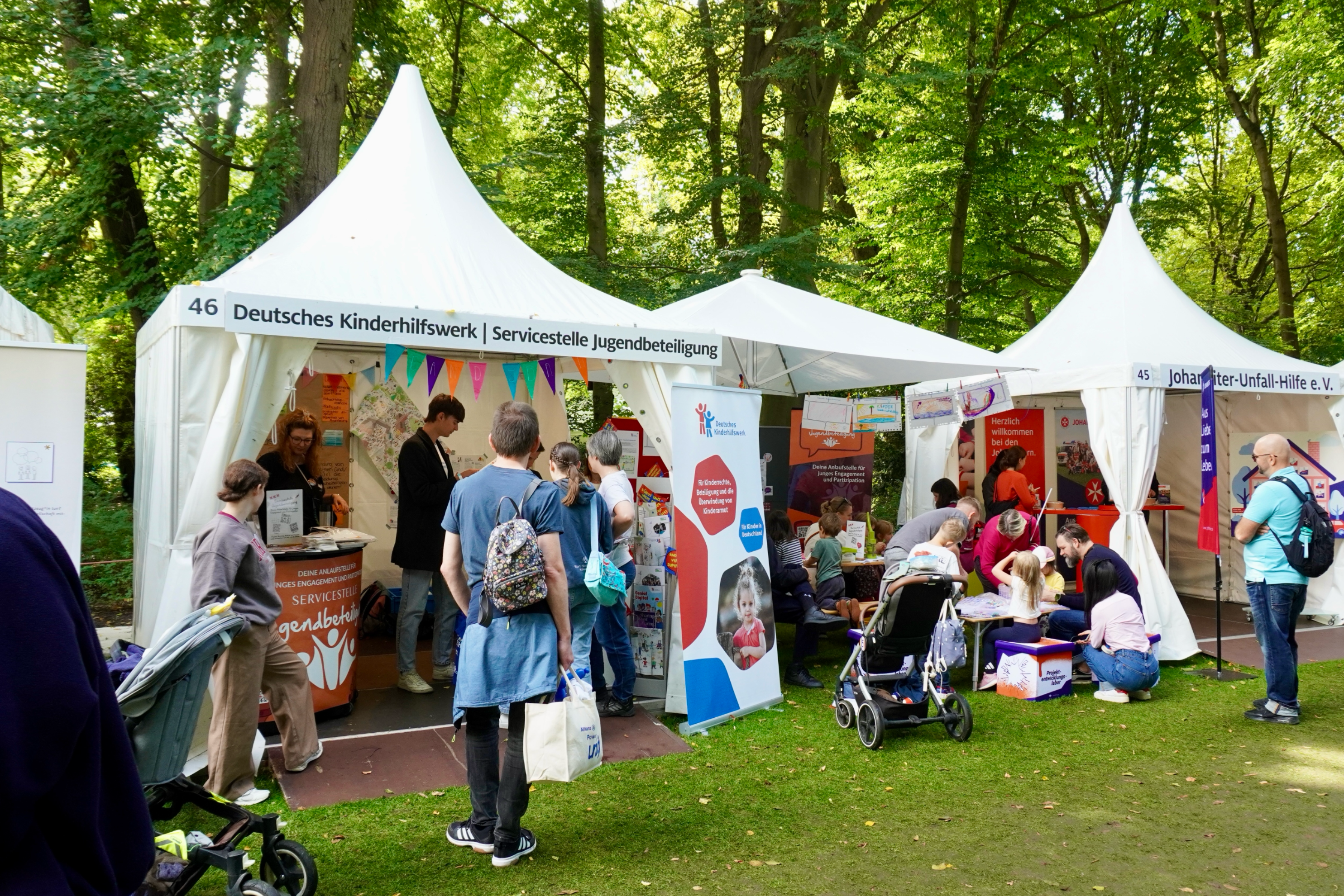 Auf dem Foto ist eine lebendige Szene beim Bürgerfest im Park von Schloss Bellevue zu sehen. Im Mittelpunkt steht ein weißes Zelt mit der Aufschrift „Deutsches Kinderhilfswerk | Servicestelle Jugendbeteiligung“. Vor und im Zelt stehen und sitzen viele Menschen, darunter Erwachsene, Jugendliche und Kinder. Links schiebt eine Person einen Kinderwagen, in der Mitte spricht jemand mit den Standbetreuer*innen. Das Zelt ist mit bunten Wimpeln geschmückt, auf den Tischen liegen Infomaterialien und Poster. Rechts daneben ist ein weiteres Zelt, vor dem Kinder mit Erwachsenen auf kleinen Hockern an Bastel- oder Malaktionen teilnehmen. Die ganze Szene spielt im Grünen, unter hohen Bäumen, was dem Bild eine offene und freundliche Atmosphäre gibt.