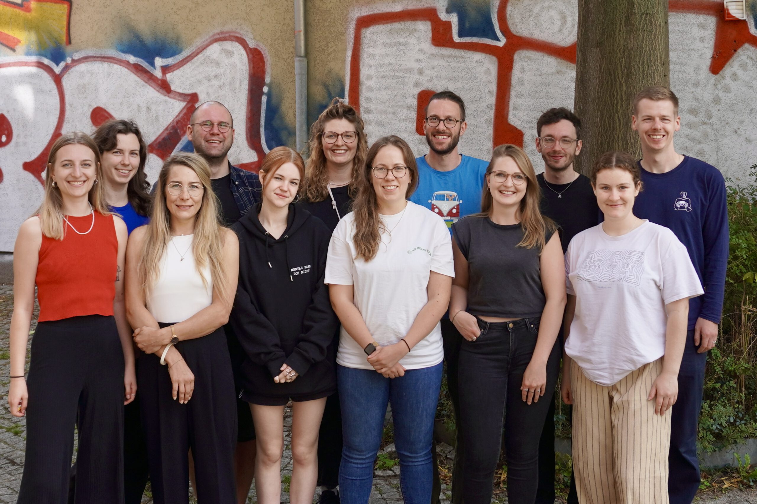 Eine Gruppe von zwölf Personen steht dicht beieinander und lächelt in die Kamera. Im Hintergrund ist eine beige Wand mit buntem Graffiti in Rot, Blau und Weiß zu sehen. Vorne stehen fünf Frauen, dahinter sieben weitere Personen, darunter Männer und Frauen. Die Stimmung wirkt fröhlich und gemeinschaftlich, es handelt sich um ein Gruppenfoto im Freien.