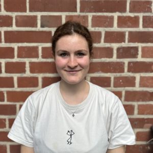 Zentriertes Brustporträt vor einer Backsteinwand mit hellen Fugen. Die Person hat zusammengebundene, lockige Haare, Sommersprossen und einen freundlichen Gesichtsausdruck. Kleidung: weißes T-Shirt mit kleinem schwarzen Figuren-Print mittig, feine silberne Kette mit kleinem Anhänger. Weiches Tageslicht, neutraler Hintergrund; die Szene wirkt natürlich und nahbar.