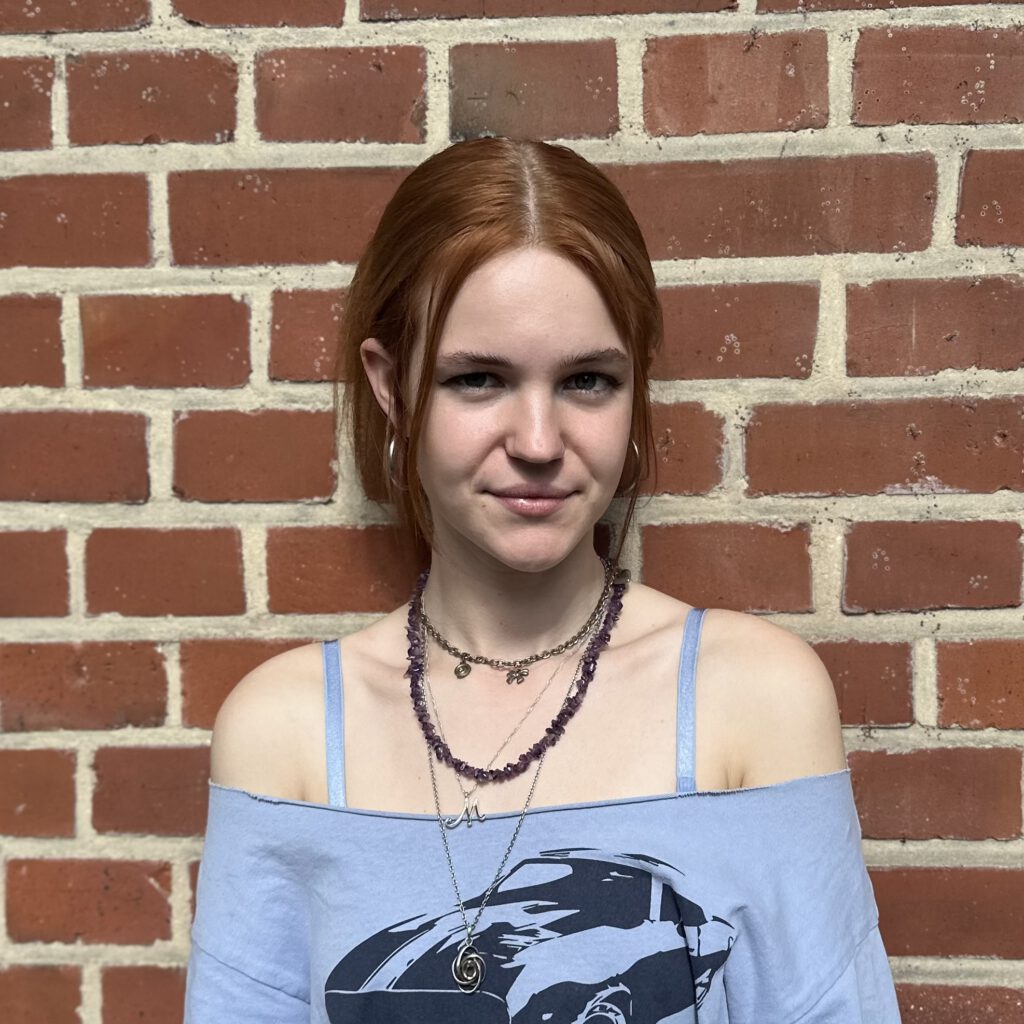 Das Bild zeigt die Geschäftsführung Lena. Sie steht vor einer mit Efeu bewachsenen Backsteinwand und lacht in die Kamera. © Servicestelle Jugendbeteiligung e.V., 2020 Anna M.