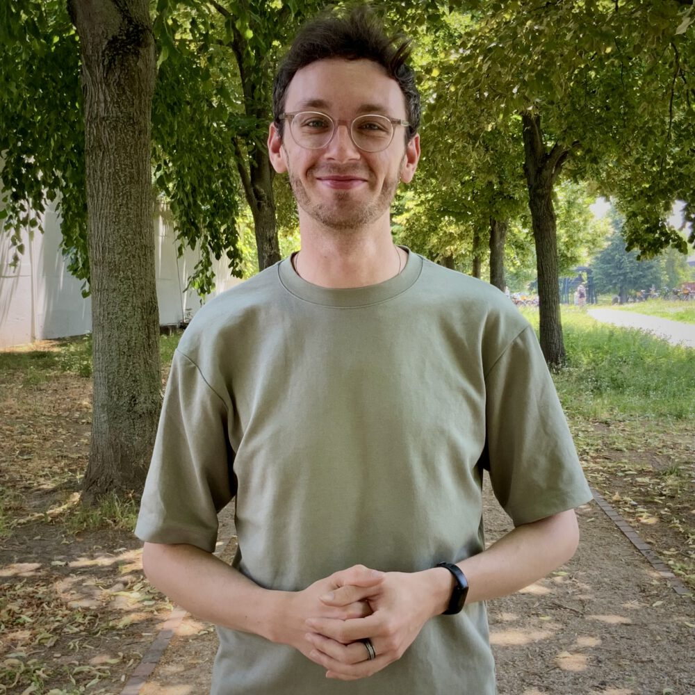 Das Bild zeigt Janis aus dem Team der Servicestelle Jugendbeteiligung. Er steht auf einem Weg mit Bäumen im Hintergrund und lacht in die Kamera. © Servicestelle Jugendbeteiligung e.V., 2025